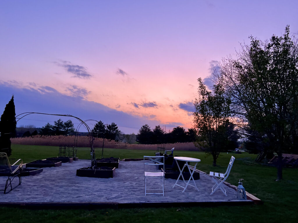view of a sunset across a garden that has a small white table and chairs in it and raised planting beds. The skies are purple and pale orange.